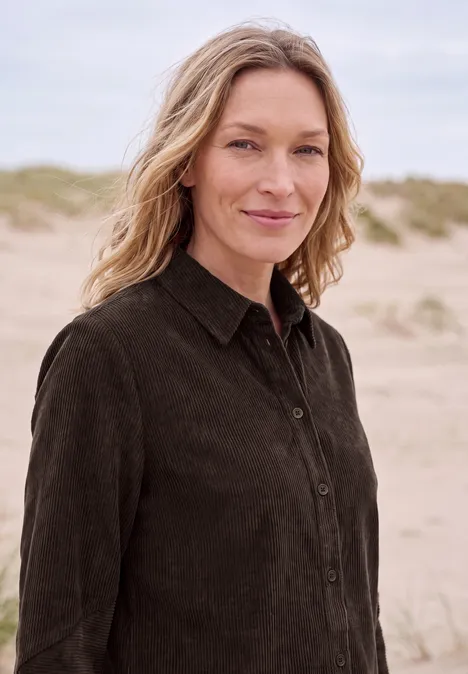 Eine Frau mit hellem Haar und dunklem Hemd steht lächelnd am Strand vor Dünen.