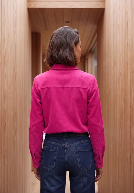 Frau steht mit Rücken zur Kamera, trägt pinke Bluse und blaue Jeans in einem hölzernen Flur.
