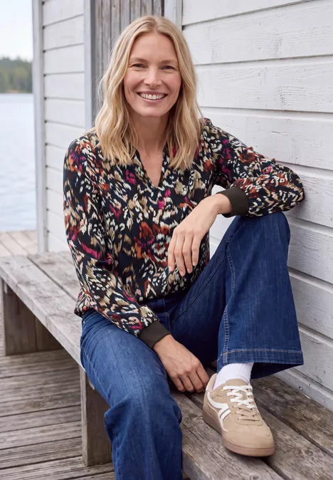 Une femme souriante en veste à motifs, jean et baskets beiges est assise sur une terrasse en bois près d'un mur blanc.