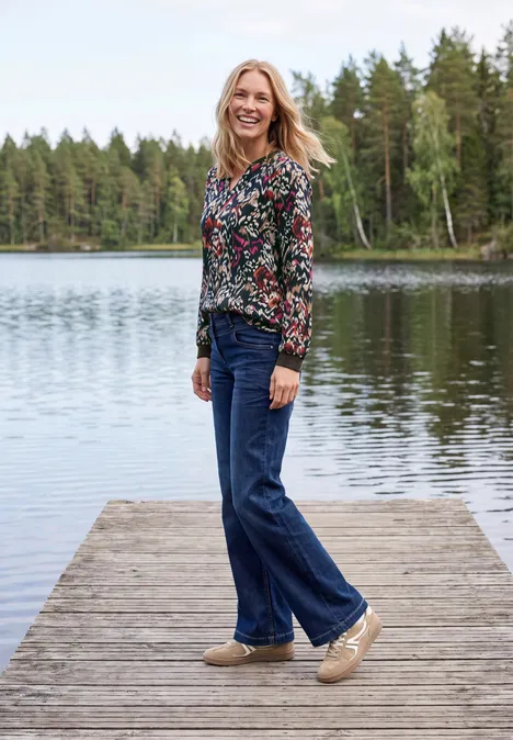 Une femme souriante est debout sur une jetée en bois au bord d’un lac paisible, forêt en arrière-plan.