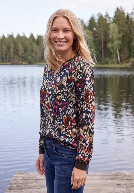 Femme souriante en chemisier coloré et jean, debout sur un ponton devant un lac et une forêt.