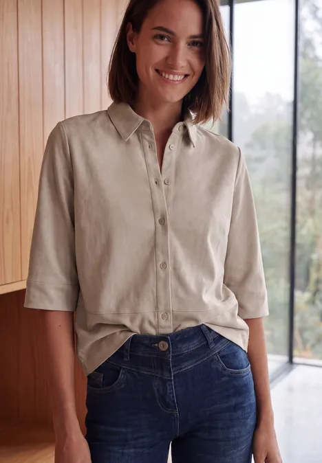 Eine lächelnde Frau in hellem Raum trägt eine beige Bluse mit ¾-Ärmeln und blaue Jeans.