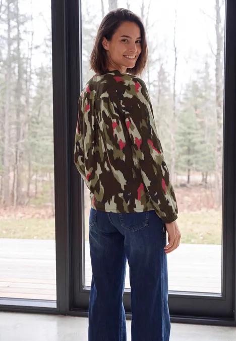 Langarm Bluse mit Rundhals und Print pink flash Eine lächelnde Frau im Camouflage-Pullover und Jeans steht vor einem großen Fenster und blickt über die Schulter.