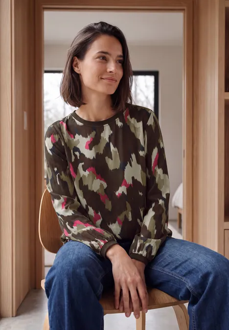 Langarm Bluse mit Rundhals und Print pink flash Frau sitzt in modernem Holzraum, trägt grünen Tarnpullover mit rosa Akzenten und blaue Jeans, schaut lächelnd zur Seite.
