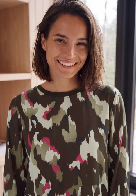 Langarm Bluse mit Rundhals und Print pink flash Junge Frau mit kurzem braunem Haar, lächelt in Camouflage-Pullover vor hellem Fenster.