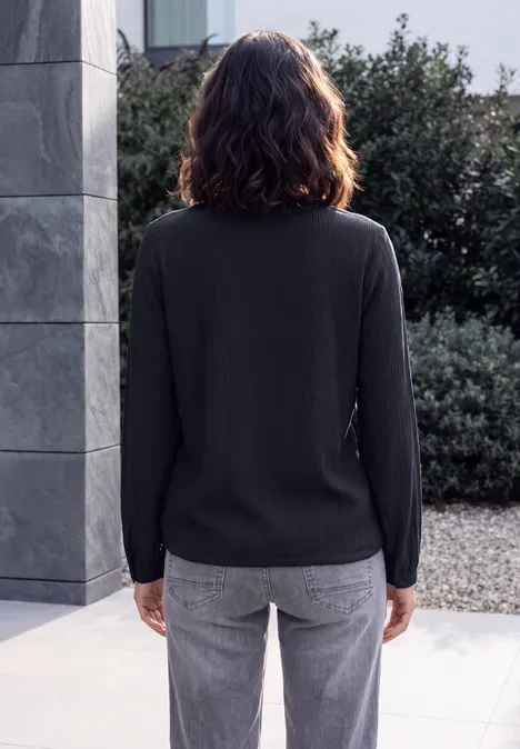 Langarm Blusenshirt mit Smokdetails urban dark blue Rückenansicht einer Frau in schwarzem Langarmshirt und grauer Jeans vor Betonwand und Gehölz.