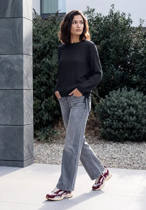 Langarm Blusenshirt mit Smokdetails urban dark blue Frau in schwarzem Pullover, grauer Jeans und Sneakern steht entspannt im Freien vor grünen Büschen.