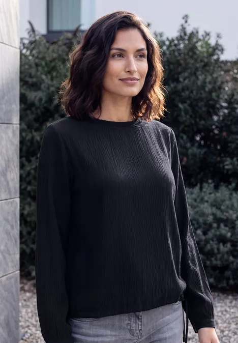 Langarm Blusenshirt mit Smokdetails urban dark blue Eine lächelnde Frau mit schulterlangem dunklem Haar trägt eine schwarze Bluse und Jeans, steht vor Hecken und einer Steinwand.