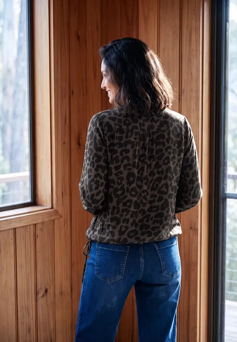 Langarm Blusenshirt mit Smokdetails truffle brown Eine Frau im Leopardenmuster-Oberteil und Jeans steht mit dem Rücken zur Kamera und blickt aus einem Holzfenster.
