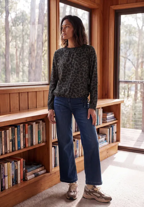 Langarm Blusenshirt mit Smokdetails truffle brown Frau in Holzhaus vor großen Fenstern, trägt Leopardenpullover, Jeans und Sneaker, steht an einem Bücherregal.