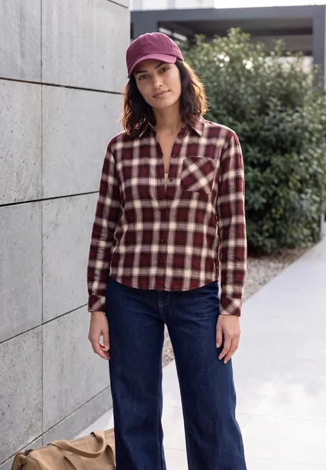 Eine Frau mit kariertem Hemd, Jeans und Baseballcap steht lässig an einer Wand im Freien.