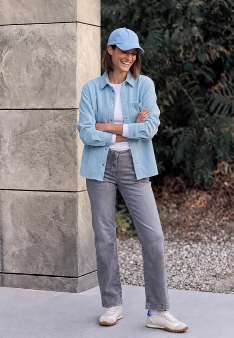 Frau in Jeanshemd, T-Shirt, grauer Hose und Cap lehnt lässig an einer Mauer im Freien.