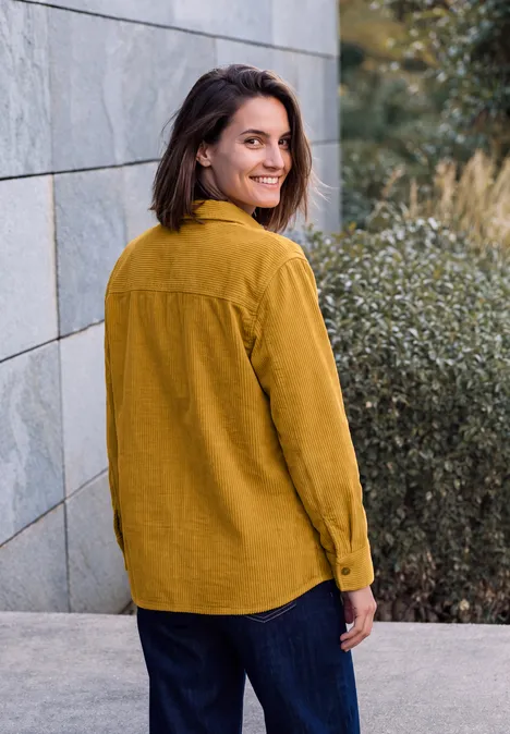 Jeune femme en chemise moutarde et pantalon sombre souriant par-dessus son épaule devant un mur de pierre.