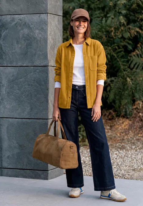 Femme en veste jaune, tee-shirt blanc, jean large et baskets, souriante, tenant un sac de voyage marron.