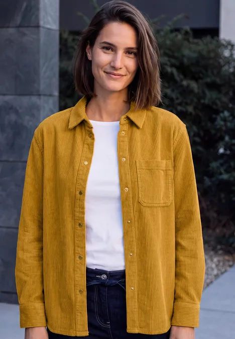 Une femme souriante se tient devant un bâtiment, portant une chemise moutarde, un t-shirt blanc et un jean foncé.