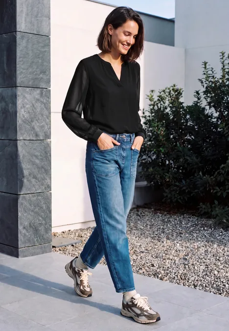 Langarm Bluse mit V-Neck und Strukturmix Black Eine junge Frau trägt schwarze Bluse, Jeans und Sneakers, steht an einer Säule im Freien und lächelt.