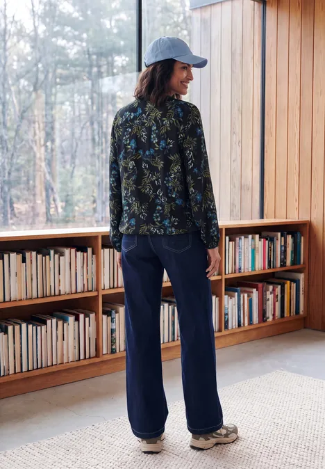 Crepe Bluse mit floralem Muster urban dark blue Frau in Cap, geblümtem Hemd und weiten Hosen steht vor einem Fensterregal voller Bücher, Blick nach draußen.