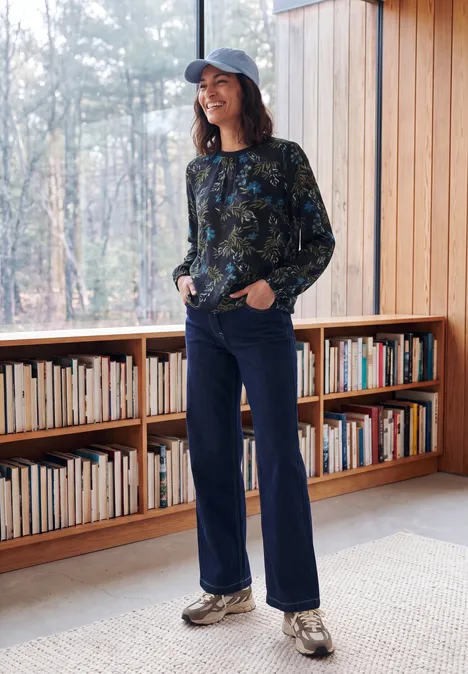 Crepe Bluse mit floralem Muster urban dark blue Eine Frau in Cap, gemustertem Pullover und Jeans steht lächelnd vor einem Bücherregal am Fenster.