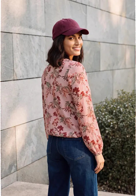 Crepe Bluse mit floralem Muster soft blossom rose Frau in Blumenbluse und Jeans, trägt eine rote Baseballkappe und lächelt vor einer Mauer mit Grün.