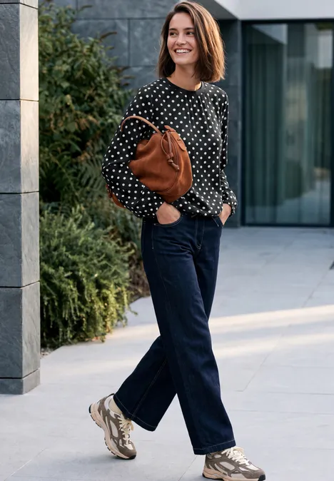 Crepe Bluse mit Punktemuster Black Lächelnde Frau in gepunkteter Bluse, Jeans und Sneakers, die eine braune Tasche an der Schulter trägt und im Freien spaziert.
