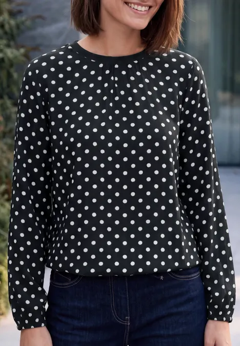 Crepe Bluse mit Punktemuster Black Frau mit schwarzer, langärmeliger Bluse mit weißen Punkten und dunkelblauer Jeans, lächelnd.