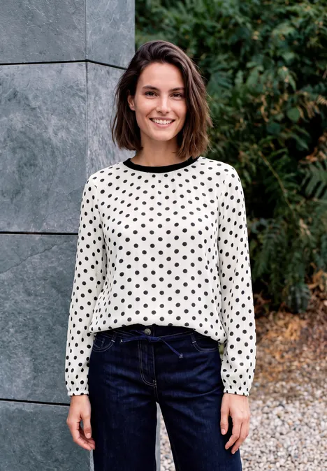 Crepe Bluse mit Punktemuster vanilla white Lächelnde Frau in weißer, schwarz gepunkteter Bluse und blauen Jeans steht draußen vor Natur und Steinwand.
