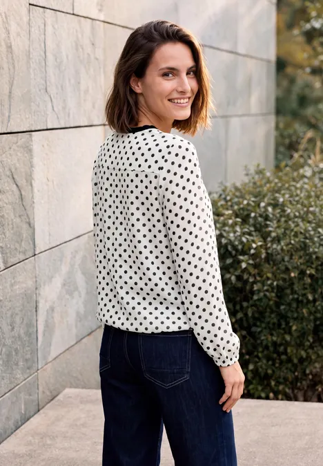 Crepe Bluse mit Punktemuster vanilla white Frau in gepunkteter Bluse und Jeans schaut lächelnd über die Schulter vor Steinmauer und Grün.