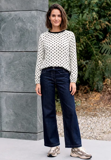 Crepe Bluse mit Punktemuster vanilla white Frau in weißem Pullover mit schwarzen Punkten, weiten Jeans und Sneakern vor grauer Wand und grünem Pflanzenhintergrund.