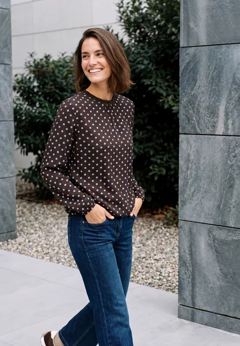 Crepe Bluse mit Punktemuster truffle brown Eine lächelnde Frau in gepunkteter Bluse und Jeans steht draußen neben steinernen Säulen.