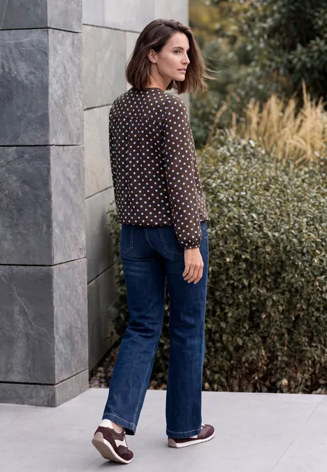 Crepe Bluse mit Punktemuster truffle brown Frau in gepunkteter Bluse und Jeans steht seitlich vor moderner Fassade und Büschen.