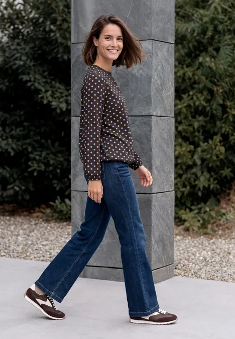 Crepe Bluse mit Punktemuster truffle brown Frau in gepunktetem Pullover und Jeans geht lächelnd an einer grauen Säule vorbei im Garten.