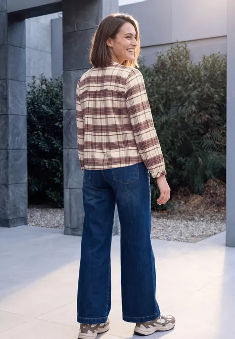 Frau steht auf einem Kiesweg vor einem modernen Gebäude, trägt kariertes Hemd, weite Jeans und Sneaker.