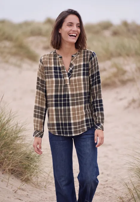Tuniek met lange mouwen en ruitjespatroon urban dark blue Een lachende vrouw in een geruit hemd en spijkerbroek staat op het strand met duinen op de achtergrond.