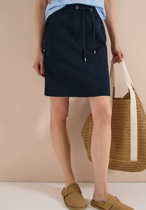 Knielanger dunkelblauer Rock, hellblaues Shirt, bequeme Slipper und geflochtene Basttasche.