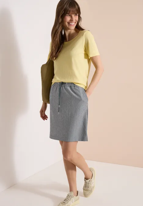 Frau in gelbem T-Shirt, grauem Rock mit Tunnelzug und Sneakers lehnt entspannt an einer Wand.