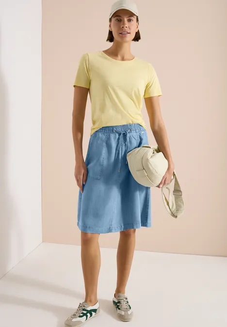 Frau in gelbem T-Shirt, leichtem Jeansrock, Sneakern, Cap und Tasche vor hellem Hintergrund.