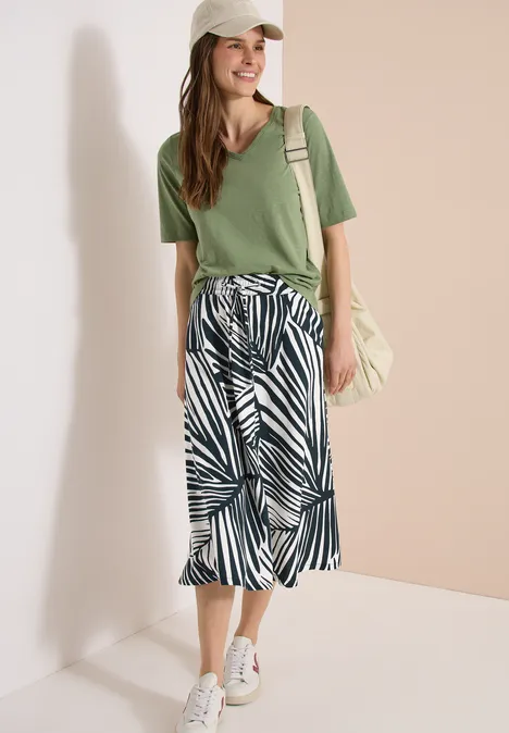 Vrouw in groen V-shirt, midi-rok met print, witte sneakers en schoudertas tegen neutrale achtergrond