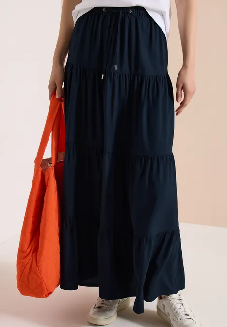 Femme portant une jupe longue bleu foncé à volants, baskets blanches et tenant un sac orange.