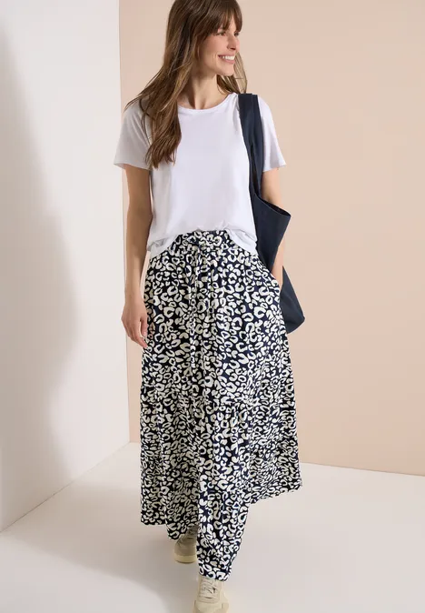 Eine Frau in weißem T-Shirt, schwarz-weißer, fließender Rock und blaue Tasche vor neutralem Hintergrund.