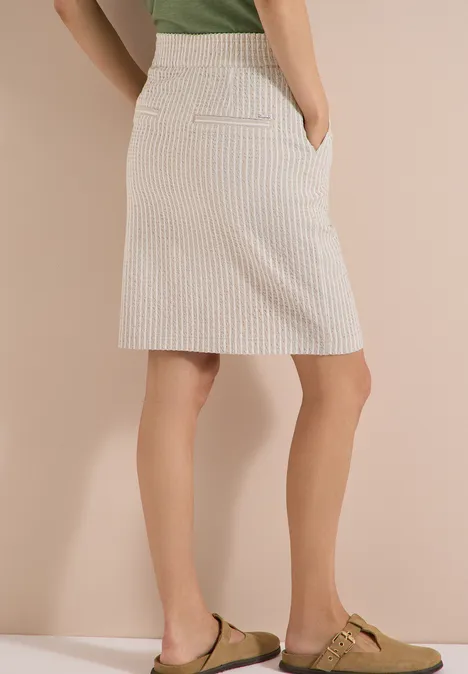 Knielanger Jogg Skirt in Seersucker-Qualität grain beige Knielanger, cremeweißer Rock mit dezenten Streifen, kombiniert mit grünem Oberteil und passenden Slippern.