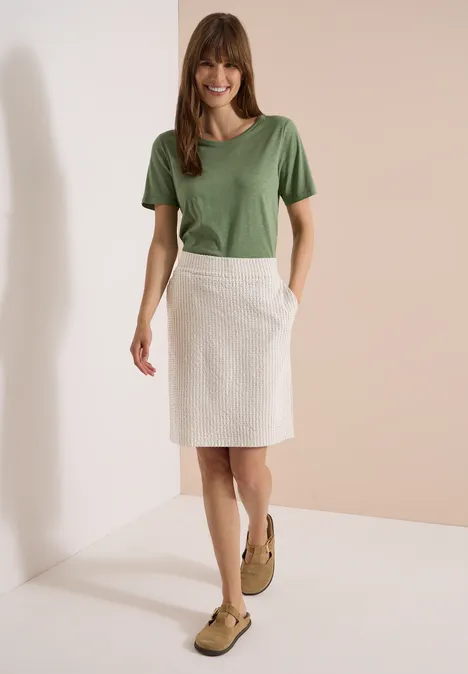 Knielanger Jogg Skirt in Seersucker-Qualität grain beige Frau in grünem T-Shirt und weißem Rock, braune Schuhe, minimalistische Kulisse.