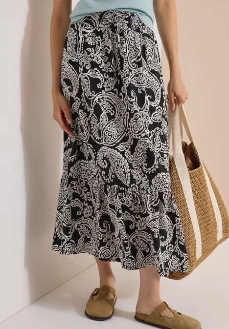Frau trägt schwarzen Maxirock mit weißem Paisley-Muster, dazu Basttasche und braune Sandalen.