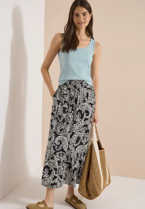 Frau in hellblauem Tanktop und schwarz-weißem Paisley-Midirock, mit geflochtener Strohtasche.