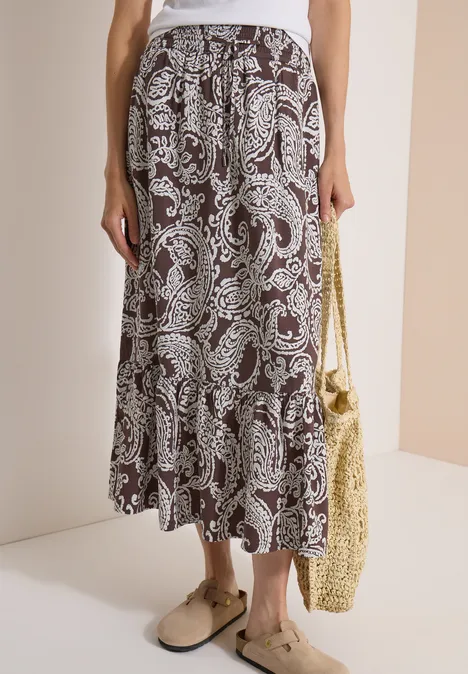 Brauner Midirock mit Paisley-Muster, Stufen, dazu beige Mules und Strohtasche.