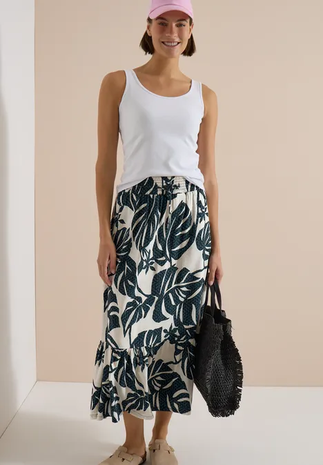 Maxi-Rock mit sommerlichem Blättermuster palm breeze green Frau trägt weißes Top und cremefarbenen Midirock mit grünem Blattmuster, dazu eine schwarze Tasche.