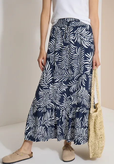 Vrouw in wit topje en lange donkerblau met wit bladmotief, strooien tas en comfortabele schoenen.