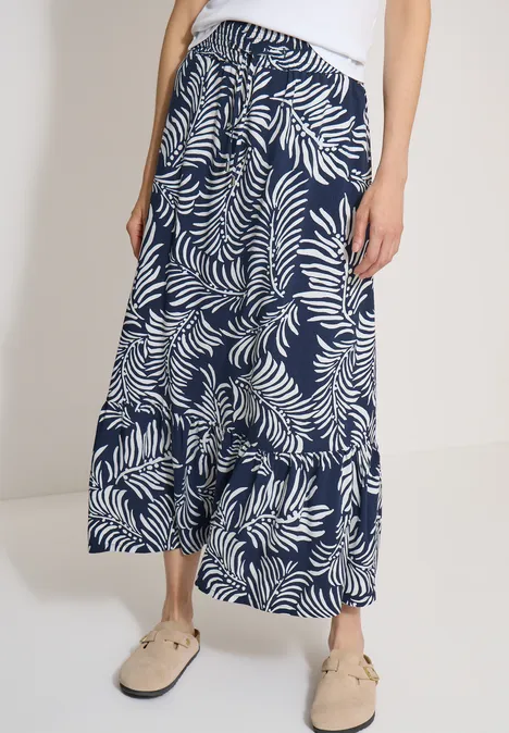 Vrouw draagt witte raffia instappers en een wijdvallende midirok met blauw-wit bladprint
