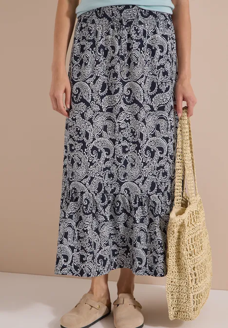 Eine Person trägt einen langen, blau-weiß gemusterten Rock im Paisley-Stil, beigefarbene Slipper und eine Basttasche.