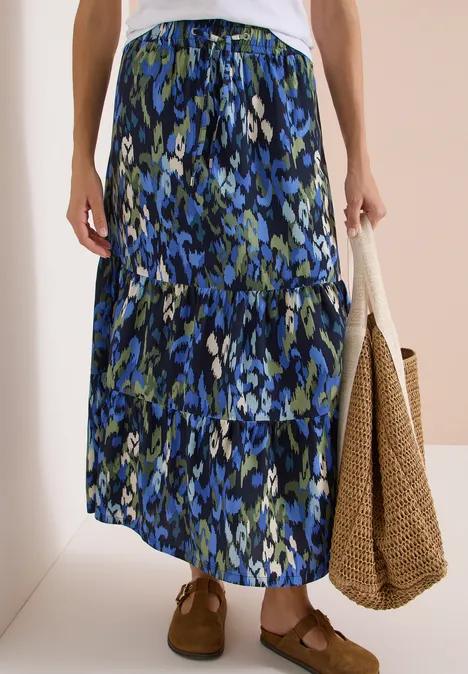 Langer Damenrock in Stufenoptik mit blau-grünem Blumenmuster, kombiniert mit geflochtener Tasche und braunen Schuhen.