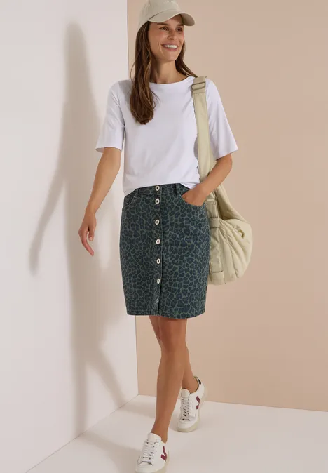 Kniehoge denimrok met luipaardprint dried olive green Vrouw in wit T-shirt, gemêleerde spijkerrok met knopen, witte sneakers en pet, nonchalant met schoudertas.